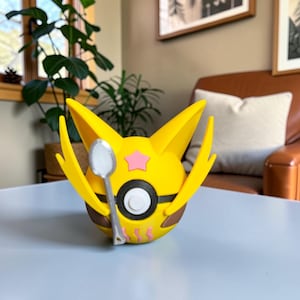 May include: A yellow and brown 3D-printed figure with a star and eye design, holding a silver spoon. The figure has large yellow ears and a black and white eye. The figure is sitting on a light blue surface.