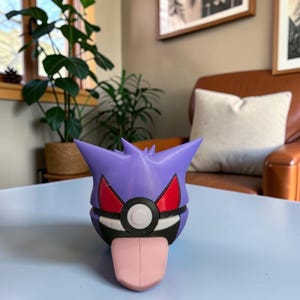 May include: A 3D-printed Gengar figure, a purple Pokémon character with red eyes, a black mouth, and a pink tongue sticking out. The figure is on a light blue surface. The Pokeforge3D logo is in the bottom right corner.