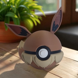 May include: A brown Eevee-themed Poké Ball figurine with large ears and a fluffy tail. The ball has a black and white center, and tan accents. The figurine is on a wooden surface with a potted plant in the background.