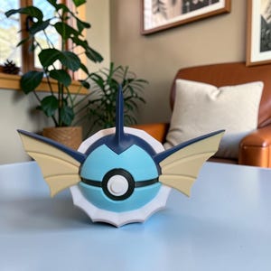 May include: A 3D-printed, stylized figure of a Pokémon character, featuring a blue, white, and tan color scheme. The figure has a spherical body with fin-like appendages and a prominent eye-like design. The figure is on a light blue surface.