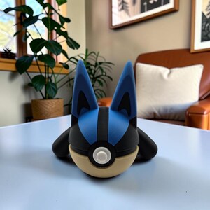 May include: A spherical figurine featuring the head of a blue and black creature with tall pointed ears, a black stripe down the center, and a tan lower section with a central white circular button, resembling a stylized character design.