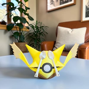 May include: A yellow, three-dimensional figure featuring two pointed ears, a brown lower section, and a central circular emblem. The figure holds two white spoons crossed in front of its face, with two wing-like appendages extending outward.