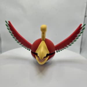 May include: A red and yellow bird-like figurine with outstretched wings. The figurine has a round body, a yellow beak, and a yellow crest. The wings are red with green and white accents. The figurine is on a white surface.