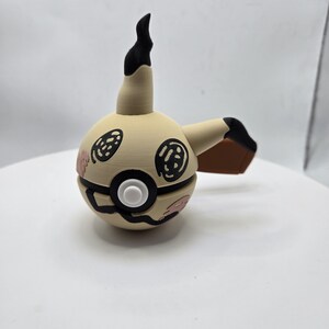 May include: A tan and black Mimikyu figure with a round body, a long neck, and a black, curved top. The figure has black eyes and a white button. The figure has a brown arm and pink accents.