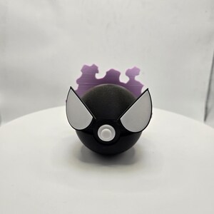 May include: A black sphere-shaped object with white and gray accents, resembling a stylized character. The object has two white, wing-like shapes on either side and a white button in the center. A purple, cloud-like shape is behind the sphere.