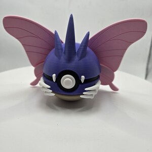 May include: A 3D-printed figurine of a creature with a purple head, white accents, and pink wings. The figurine has a round body with a large eye and a white circle in the center. The wings are spread out, and the figurine is on a white surface.