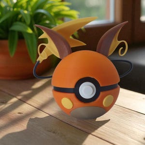 May include: An orange and black spherical toy with yellow accents and a white circle. The toy has brown and yellow wing-like features and is resting on a wooden surface. A potted plant is in the background.