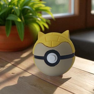 May include: A yellow and tan novelty item shaped like a cartoon character's head, with a black stripe and white button detail. The item is sitting on a wooden surface, with a potted plant in the background.