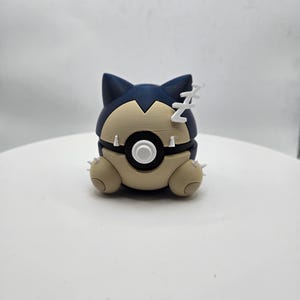 May include: A 3D-printed Snorlax figure in a Poké Ball design. The figure features a navy blue head with pointed ears and a beige body. The figure has a white button and a black circle in the center. White 'Z's are above the head.