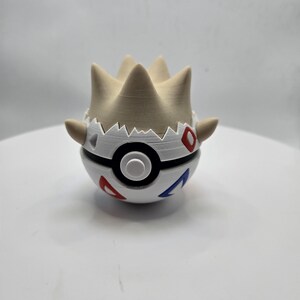 May include: A 3D-printed Togepi figure, a Pokémon character, in a white and black Poké Ball design. The figure has red and blue accents, with a beige crown-like top. The figure is on a white surface.