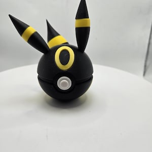 May include: A black and yellow figurine of a creature with three pointed ears. The figurine has a round body with a white button and a yellow ring around a black eye. The ears are black with yellow stripes.