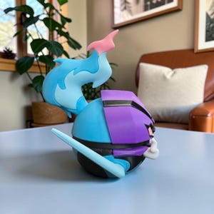 May include: A spherical character figurine featuring a bright blue flame-like crest on top, a purple helmet-like section, and a black base. The figure has a light blue pointed protrusion extending from the side and a small white hand-like detail.