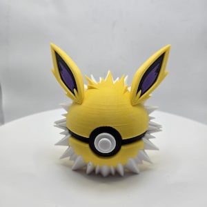 May include: A yellow and white Jolteon-themed Poké Ball. The ball features a yellow body with white spikes, black and white accents, and two yellow ears with purple details. A unique collectible.