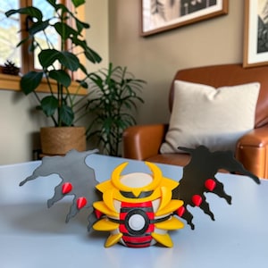 May include: A 3D-printed, yellow, red, and black figure with wing-like extensions. The figure has a circular design in the center and is set on a light blue surface. The figure is a Pokemon character.