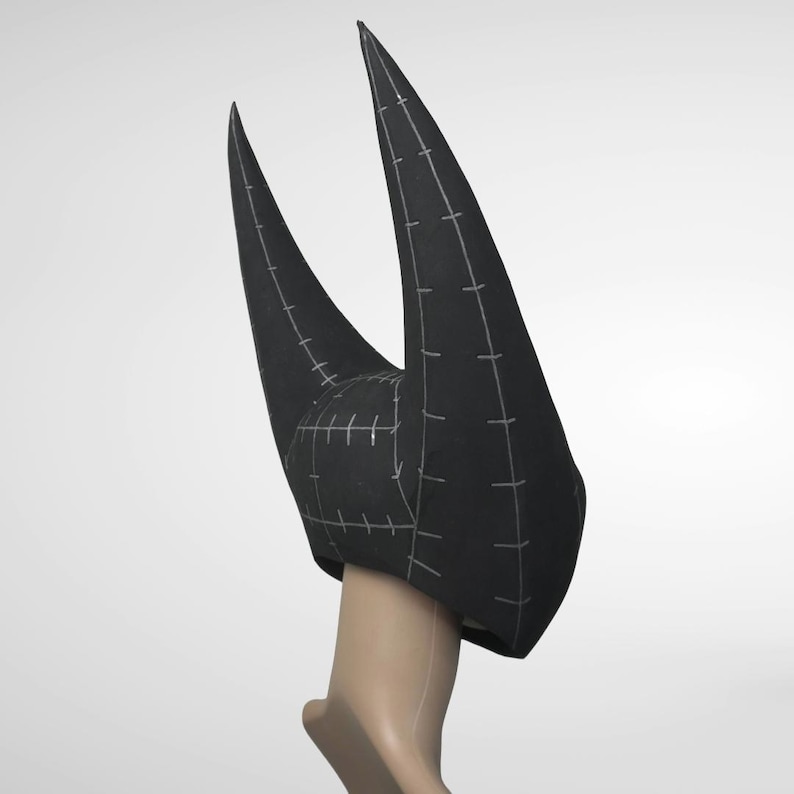 May include: A black, horn-shaped headpiece with a pointed design. The headpiece is made of a dark fabric and features white stitching lines. The horns are long and curve upwards. The headpiece is on a mannequin head.