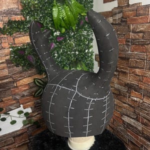 May include: A black, handmade headpiece with two curved horns and a rounded base. White stitching lines and grid patterns are visible across the surface. The headpiece is displayed on a white mannequin head.