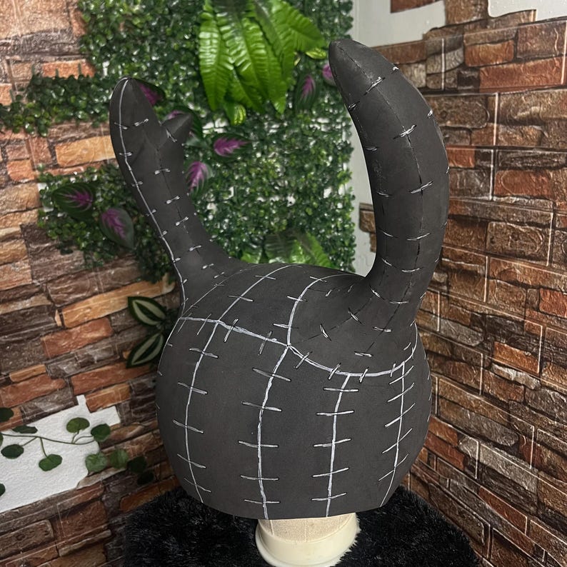 May include: A black, handmade headpiece with two curved horns and a rounded base. White stitching lines and grid patterns are visible across the surface. The headpiece is displayed on a white mannequin head.