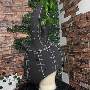 May include: A black, handmade hat with two tall, curved protrusions, possibly bunny ears. White stitching lines are visible across the surface, indicating a pattern or design. The hat is displayed on a mannequin head against a backdrop of brick and greenery.