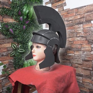 May include: A black Roman helmet with a tall crest, displayed on a mannequin head with a red cloth. The helmet has ear guards and is decorated with white lines. The background features a brick wall and greenery.