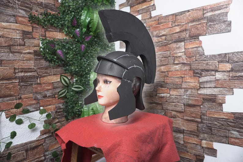 May include: A black Roman helmet with a tall crest, displayed on a mannequin head with a red cloth. The helmet has ear guards and is decorated with white lines. The background features a brick wall and greenery.