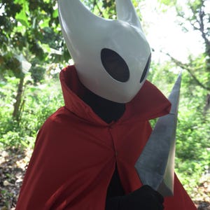May include: A white mask with two horns and large eye openings, worn with a red cape and black gloves. The person is holding a large, gray, blade-like weapon. The costume is set against a backdrop of green foliage.
