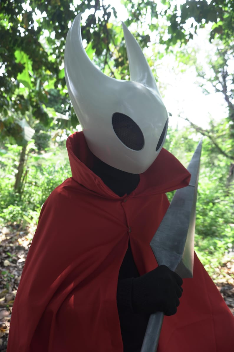 May include: A white mask with two horns and large eye openings, worn with a red cape and black gloves. The person is holding a large, gray, blade-like weapon. The costume is set against a backdrop of green foliage.