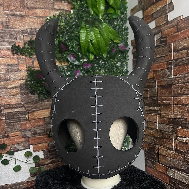 May include: A black mask with large, curved horns and stitched detailing. The mask has two large eye holes and is set against a backdrop of greenery and a brick wall. The mask is designed to be worn.