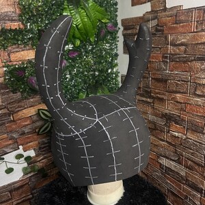 May include: A black, horn-shaped headpiece with white stitching details. The headpiece is made of a foam-like material and is designed to sit on a head form. The horns curve upwards and outwards, suggesting a costume or cosplay item.