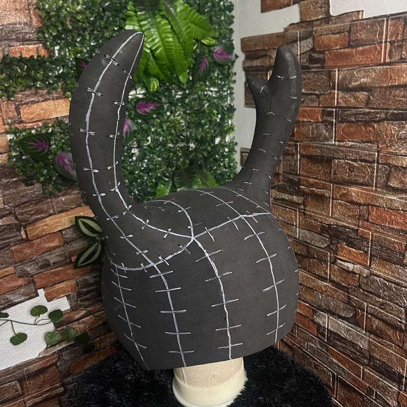 May include: A black, horn-shaped headpiece with white stitching details. The headpiece is made of a foam-like material and is designed to sit on a head form. The horns curve upwards and outwards, suggesting a costume or cosplay item.