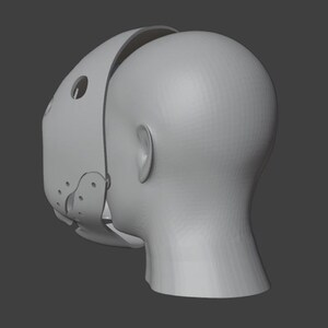 Op de afbeelding: Een lichtgrijze, 3D-geprinte muilkorfmasker met een gebogen ontwerp, met cirkelvormige en ovale openingen. Het masker is gedeeltelijk bevestigd aan een 3D-geprinte kop, met het oor en de nek.