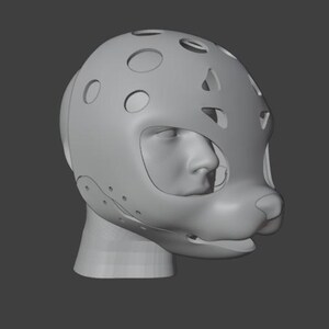 Op de afbeelding: Een grijze, 3D-gemodelleerde hoofdbedekking met een gestileerd dierengezichtontwerp. Het masker heeft meerdere cirkelvormige en ovale uitsparingen, met openingen voor de ogen en mond. Het ontwerp omvat een neus en een afgeronde snuit.