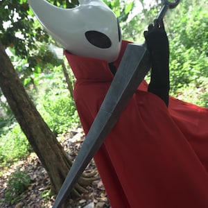 May include: A cosplay costume featuring a white horned mask with black eye holes, a red cape, and a large, gray, pointed sword. The costume is set against a backdrop of green foliage and trees.
