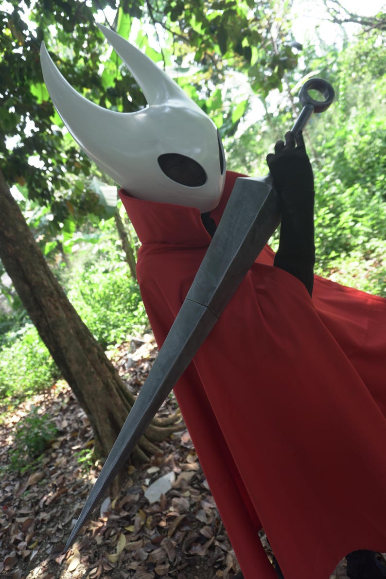 May include: A cosplay costume featuring a white horned mask with black eye holes, a red cape, and a large, gray, pointed sword. The costume is set against a backdrop of green foliage and trees.