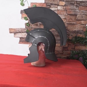 May include: A black helmet with a tall, curved crest, possibly for cosplay or costume use. The helmet is placed on a mannequin head, with white markings and a red surface in the foreground. The background features a brick wall and greenery.