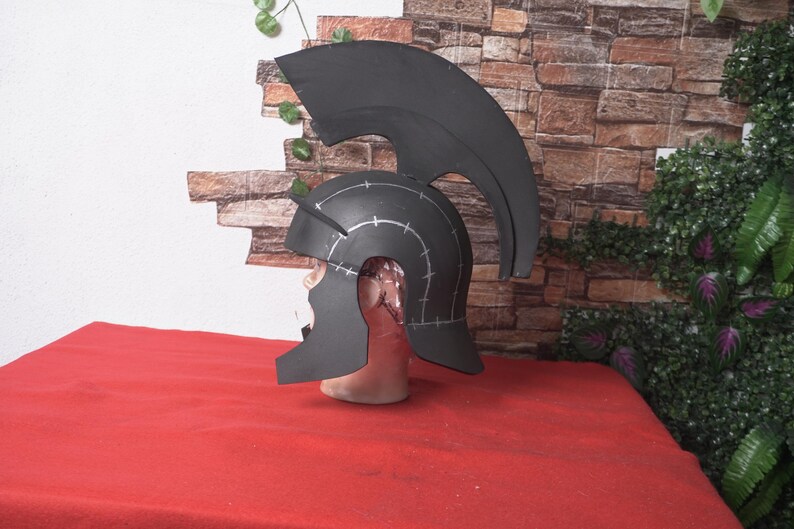 May include: A black helmet with a tall, curved crest, possibly for cosplay or costume use. The helmet is placed on a mannequin head, with white markings and a red surface in the foreground. The background features a brick wall and greenery.