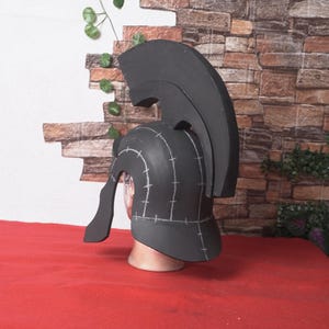 May include: A black helmet with a tall crest, reminiscent of ancient Greek or Roman designs. The helmet is displayed on a mannequin head, set against a brick wall and red surface. The helmet has white markings.