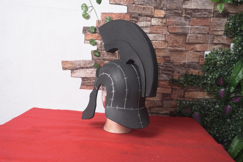 May include: A black helmet with a tall crest, reminiscent of ancient Greek or Roman designs. The helmet is displayed on a mannequin head, set against a brick wall and red surface. The helmet has white markings.