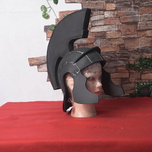 May include: A black foam Roman helmet with a tall crest, displayed on a mannequin head. The helmet has a detailed design with visible construction lines. The background features a brick wall and greenery, with the helmet resting on a red surface.