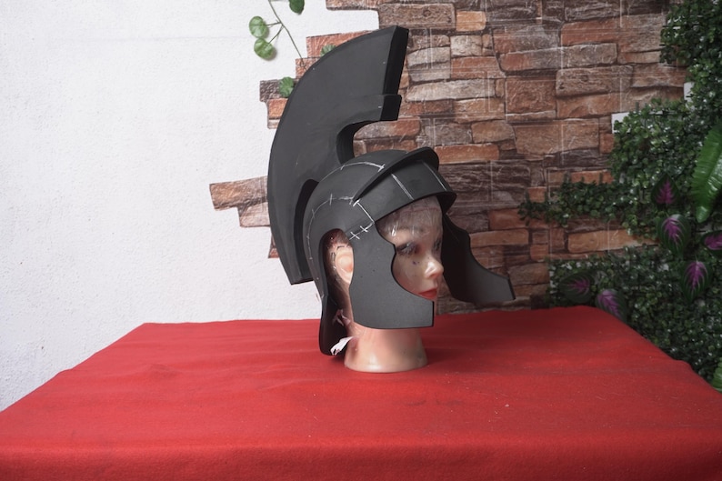 May include: A black foam Roman helmet with a tall crest, displayed on a mannequin head. The helmet has a detailed design with visible construction lines. The background features a brick wall and greenery, with the helmet resting on a red surface.