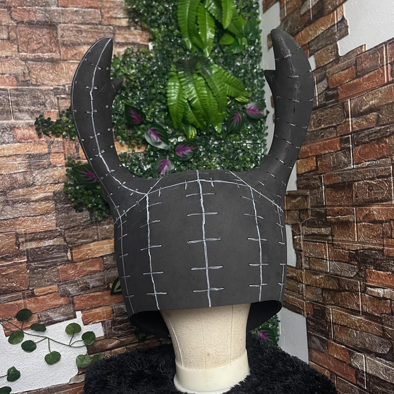 May include: A black horned helmet with white grid lines and stitching details. The helmet has two large, curved horns extending upwards. The design suggests a costume or cosplay item, possibly for a fantasy or mythical character.
