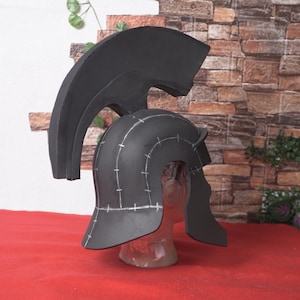 May include: A black Spartan helmet with a large, curved crest. The helmet has a rounded crown and cheek guards, with white markings. It is displayed on a clear mannequin head against a brick wall.