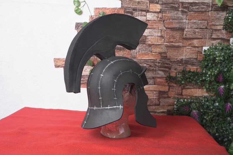 May include: A black Spartan helmet with a large, curved crest. The helmet has a rounded crown and cheek guards, with white markings. It is displayed on a clear mannequin head against a brick wall.