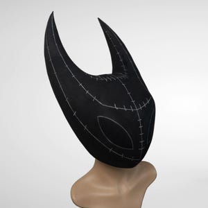 May include: A black mask with two curved horns and a stitched design. The mask has a stylized face shape with eye openings. The mask is displayed on a mannequin head.