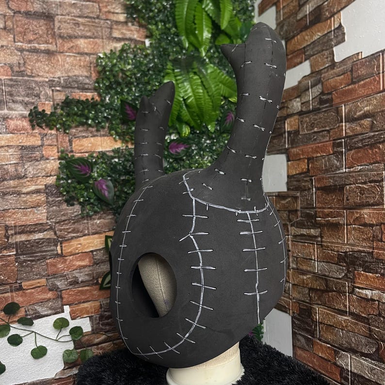 May include: A black mask with two tall, curved protrusions and a large oval opening for the face. The mask is constructed from a dark material and features white stitching details throughout. The mask is designed to be worn.