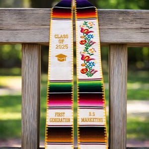 Puede incluir: Una estola de graduación con rayas tejidas coloridas y bordados florales. La estola tiene el texto "CLASS OF 2025", un gráfico de gorro de graduación y las palabras "FIRST GENERATION" y "B.S MARKETING". La estola está colgada en un banco de madera.