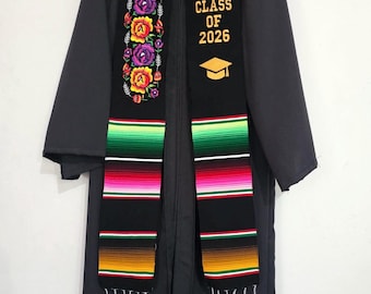 May include: A black graduation gown with a colorful serape-style stole. The stole features the text "CLASS OF 2026" with a mortarboard graphic, and is adorned with floral embroidery. The stole has a striped pattern in red, green, and yellow.
