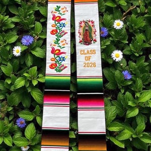 Puede incluir: Una estola de graduación blanca con rayas tejidas coloridas y bordados florales. Un lado presenta una imagen de la Virgen María y el texto "CLASS OF 2026" en letras doradas. La estola tiene flecos blancos.