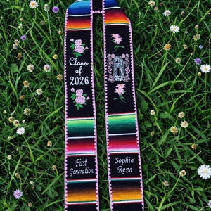 May include: A black graduation sash with vibrant striped detailing and pink edging. It features embroidered text reading "Class of 2026", floral motifs, and the phrases "First Generation" and "Sophia Reza". The sash is laid on a bed of green grass and small white flowers.