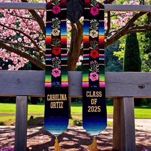 以下が含まれることがあります： カラフルな刺繍の花柄と、金色の文字で「CAROLINA ORTIZ」と「CLASS OF 2025」の文字が入った卒業ストールが2つ。ストールは黒を基調とし、グラデーションの青と緑の裾と金色のタッセルが付いています。