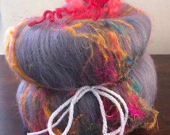 May include: A two-layered ball of yarn, primarily gray, with vibrant streaks of yellow, orange, pink, and blue. The top layer has a tuft of bright red yarn. A white string is tied around the middle. Ideal for knitting and crochet projects.
